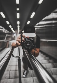 A person wearing a cap is holding a camera, capturing a shot while leaning down on a modern, sleek staircase with metallic railings. The perspective creates a dynamic, symmetrical view along the lines of the railings and the tiles, with ceiling lights adding depth.
