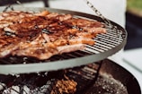 A barbecue grill with sizzling meat pieces cooking over an open flame. The grill is circular and elevated, displaying a variety of browned and caramelized meats against a backdrop of ashes and glowing embers.