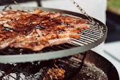 A barbecue grill with sizzling meat pieces cooking over an open flame. The grill is circular and elevated, displaying a variety of browned and caramelized meats against a backdrop of ashes and glowing embers.