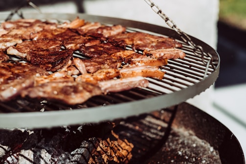 A barbecue grill with sizzling meat pieces cooking over an open flame. The grill is circular and elevated, displaying a variety of browned and caramelized meats against a backdrop of ashes and glowing embers.