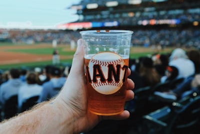 person holding clear plastic cup with beer giants teams background