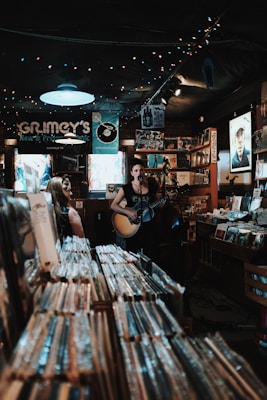 A musician is performing with a guitar in a dimly lit record store, surrounded by shelves filled with vinyl records and music memorabilia. The store is decorated with colorful string lights and posters. A small crowd is gathered to listen.