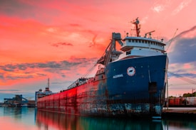 A large cargo ship docked at a harbor during sunset, with vibrant orange and pink hues filling the sky. The ship's hull is painted black and red, and it reflects on the calm water. Industrial structures and containers are visible in the background.