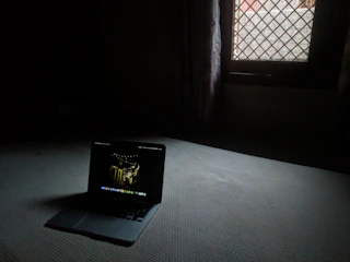 Dark room with a laptop showing a shadowy figure and eerie urban scenes.