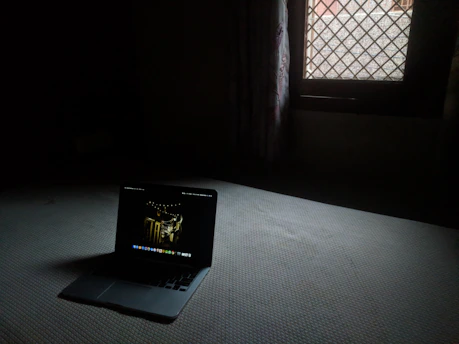 Dark room with a laptop showing a shadowy figure and eerie urban scenes.