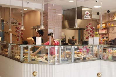 A bakery display inside a shop with a variety of pastries and sweets. Two employees are attending to the products, with one arranging items and the other interacting with a customer. The interior is decorated with light colors and glass display cases. Lollipop decorations hang from above, adding a festive touch.