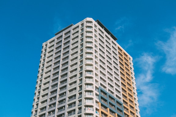 A tall, modern apartment building with many floors and a mix of grey and light brown facade panels, set against a clear blue sky. The windows are lined in rows, typical of a residential high-rise.