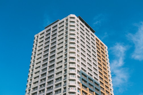 A tall, modern apartment building with many floors and a mix of grey and light brown facade panels, set against a clear blue sky. The windows are lined in rows, typical of a residential high-rise.