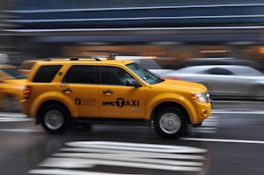 A yellow NYC taxi is captured in motion on a city street. The background is blurred, highlighting the speed of the taxi as it moves along the wet road. Multiple lanes of traffic are visible with blurred vehicles, emphasizing urban hustle.