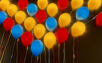 A colorful arrangement of illuminated balloons in red, yellow, and blue, affixed to a wall. The balloons create a vibrant and festive atmosphere, with strings attached to each balloon.
