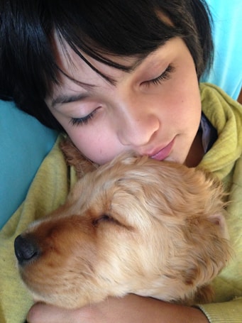 A young child with dark hair is sleeping peacefully, cuddling a sleeping light brown puppy. The child appears to be relaxed, wearing a yellow sweatshirt. The scene conveys a sense of warmth and comfort with both the child and the puppy in close embrace.
