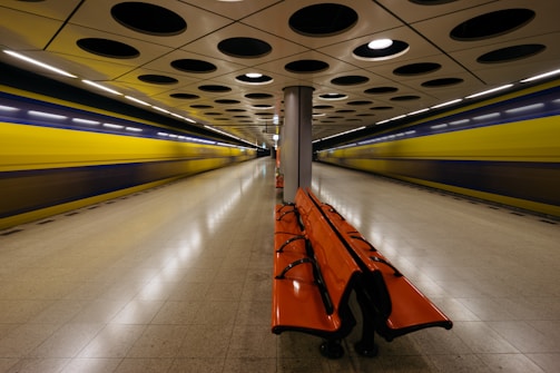 A subway station platform with a sleek, modern design features a row of bright orange benches positioned centrally. The ceiling, adorned with a pattern of circular black recesses, complements the polished floor tiles. On either side of the platform, two blurred yellow trains convey a sense of motion and speed.
