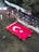 A large group of people is gathered in an outdoor setting with a focus on a sizable Turkish flag being held by several individuals wearing red shirts. The assembly takes place on a grassy area surrounded by banners and spectators seated in a shaded zone. The environment is festive, with national flags adorning the scene.