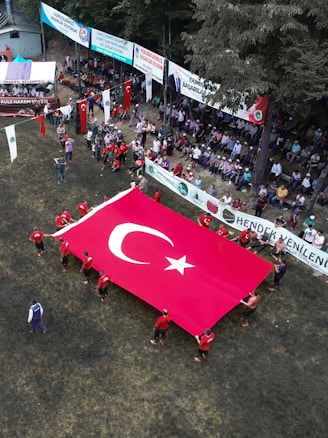A large group of people is gathered in an outdoor setting with a focus on a sizable Turkish flag being held by several individuals wearing red shirts. The assembly takes place on a grassy area surrounded by banners and spectators seated in a shaded zone. The environment is festive, with national flags adorning the scene.