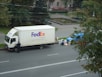 A large delivery truck and a smaller blue van are parked on the side of a road next to a park. A person is standing near the truck's door. Several packages and boxes are placed on the ground between the vehicles. The surrounding area features green trees and a small water fountain within a park setting.