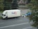 A large delivery truck and a smaller blue van are parked on the side of a road next to a park. A person is standing near the truck's door. Several packages and boxes are placed on the ground between the vehicles. The surrounding area features green trees and a small water fountain within a park setting.