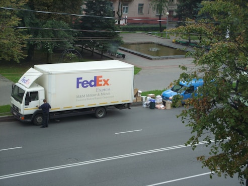 A large delivery truck and a smaller blue van are parked on the side of a road next to a park. A person is standing near the truck's door. Several packages and boxes are placed on the ground between the vehicles. The surrounding area features green trees and a small water fountain within a park setting.