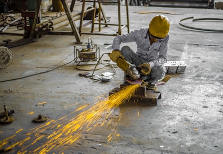 A person wearing a yellow hard hat and protective gloves is using an angle grinder, producing bright orange sparks. The scene is set in an industrial environment with a concrete floor and scattered equipment.