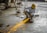 A person wearing a yellow hard hat and protective gloves is using an angle grinder, producing bright orange sparks. The scene is set in an industrial environment with a concrete floor and scattered equipment.