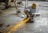 A person wearing a yellow hard hat and protective gloves is using an angle grinder, producing bright orange sparks. The scene is set in an industrial environment with a concrete floor and scattered equipment.