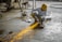 A person wearing a yellow hard hat and protective gloves is using an angle grinder, producing bright orange sparks. The scene is set in an industrial environment with a concrete floor and scattered equipment.