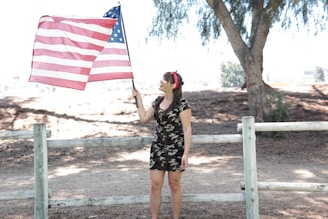 A proud woman wearing a First Ladies patriotic t-shirt, standing with a waving American flag behind her.