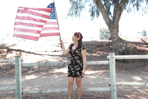 A proud woman wearing a First Ladies patriotic t-shirt, standing with a waving American flag behind her.
