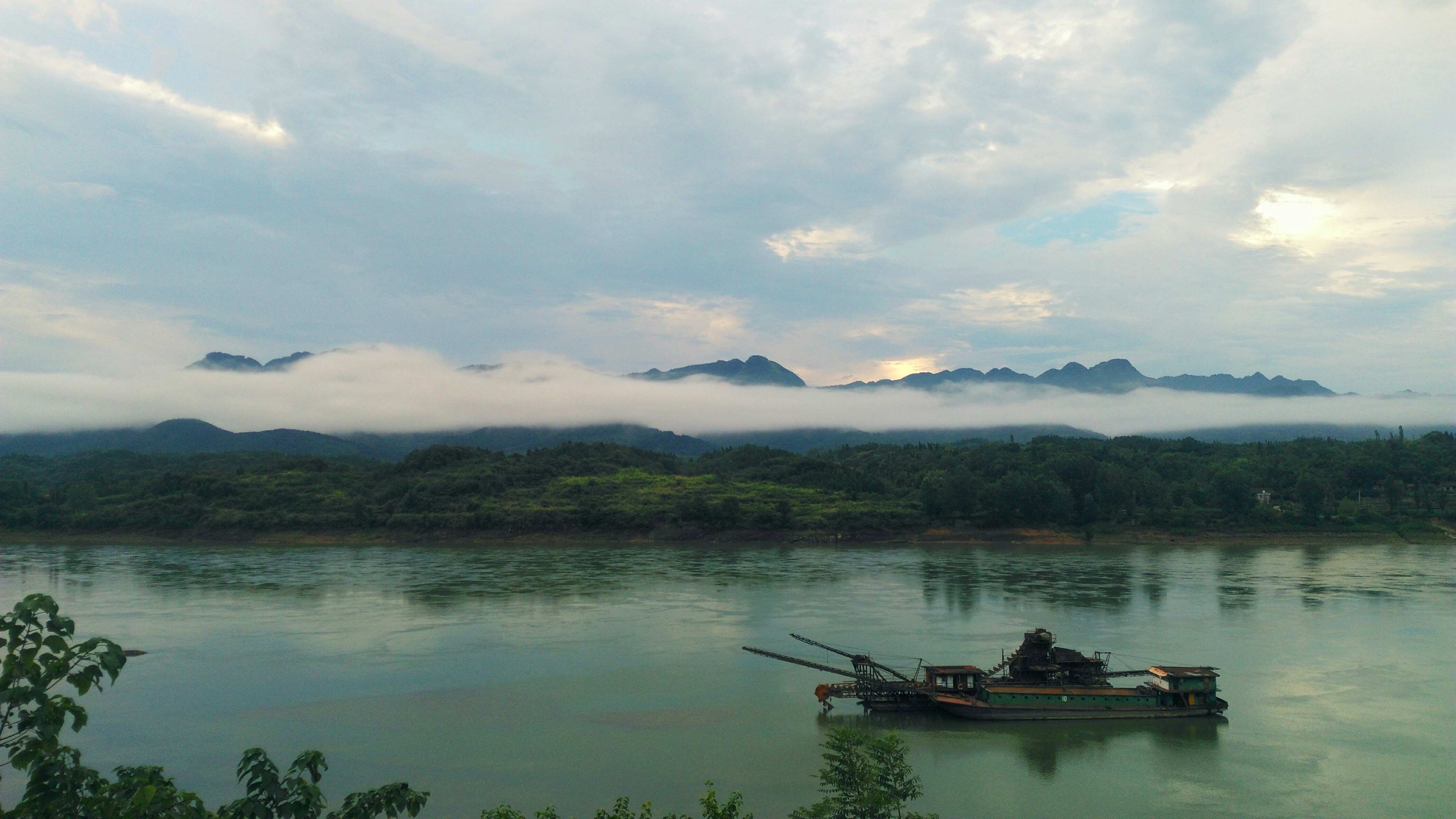 Dredging vessel operating in a wide river channel, murky brown water, industrial port infrastructure in background, overcast sky