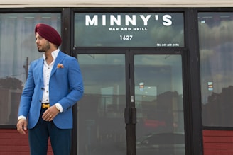 A man wearing a blue blazer, white shirt, and red turban stands outside a closed establishment named Minny's Bar and Grill. The man has a confident stance with one hand in his pocket and the other adjusting his blazer. The glass door reflects the cloudy sky and nearby structures.