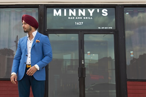 A man wearing a blue blazer, white shirt, and red turban stands outside a closed establishment named Minny's Bar and Grill. The man has a confident stance with one hand in his pocket and the other adjusting his blazer. The glass door reflects the cloudy sky and nearby structures.