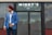 A man wearing a blue blazer, white shirt, and red turban stands outside a closed establishment named Minny's Bar and Grill. The man has a confident stance with one hand in his pocket and the other adjusting his blazer. The glass door reflects the cloudy sky and nearby structures.