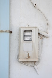 A weathered, white electrical meter mounted on a textured white wall with visible rust and discoloration on the edges. The meter is connected to several white cables that are slightly aged and curved around the device. Adjacent to the meter is a pale blue strip which appears to be a part of the wall or a shutter.