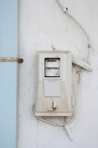 A weathered, white electrical meter mounted on a textured white wall with visible rust and discoloration on the edges. The meter is connected to several white cables that are slightly aged and curved around the device. Adjacent to the meter is a pale blue strip which appears to be a part of the wall or a shutter.
