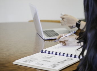 Two individuals are seated at a wooden table, one using a laptop while the other writes in a notebook with printed images. The scene appears to be a professional or educational setting.