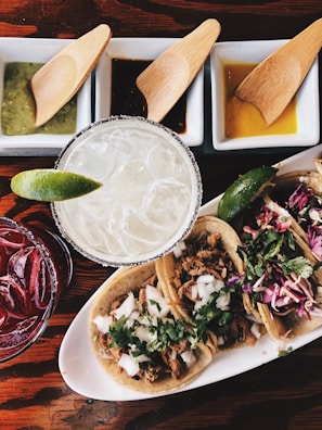 Three tacos on a white plate are accompanied by two drinks, one with a salted rim and a lime wedge, and another reddish drink. There are also three small dishes of different sauces or dips each with a wooden spoon.