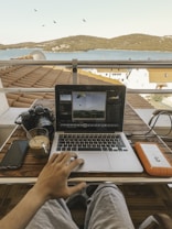 A person is seated outdoors on a balcony overlooking a serene sea view with distant hills. In front of them, there is a laptop displaying an image editing software, with a hand interacting with the trackpad. On the table, there is a camera, a smartphone, a glass of coffee, and an orange external hard drive. Birds are flying in the sky above.