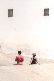 Two individuals are standing against a large, light-colored wall with two small, barred windows. One person wears a red shirt, and the other wears a black top. They appear to be facing the wall, with sunlight casting shadows on the ground.