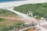 A scenic beach area with sandy shores and a clear pathway leading to the sea. The foreground features numerous bicycles parked next to a paved walkway bordered by grassy dunes. People are scattered throughout, enjoying the sunny day. Colorful beach huts and umbrellas are visible in the distance near the shoreline.