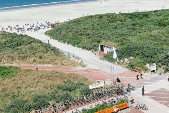 A scenic beach area with sandy shores and a clear pathway leading to the sea. The foreground features numerous bicycles parked next to a paved walkway bordered by grassy dunes. People are scattered throughout, enjoying the sunny day. Colorful beach huts and umbrellas are visible in the distance near the shoreline.