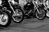 Several motorcycles are lined up on a cracked pavement. The foreground shows detailed views of the front wheels and chrome parts of the bikes. Riders with casual attire, such as jeans and t-shirts, are partially visible. The image is in black and white, emphasizing contrast and texture.