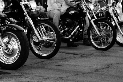 Several motorcycles are lined up on a cracked pavement. The foreground shows detailed views of the front wheels and chrome parts of the bikes. Riders with casual attire, such as jeans and t-shirts, are partially visible. The image is in black and white, emphasizing contrast and texture.