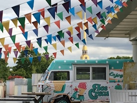 A colorful outdoor scene features an ice cream truck with polka dot patterns and playful designs. Above it, strings of vibrant triangular flags in various colors add to a festive atmosphere. In the background, a lush green area and a clock tower are visible, providing a serene backdrop.