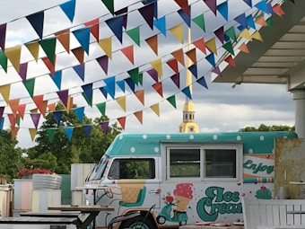 A colorful outdoor scene features an ice cream truck with polka dot patterns and playful designs. Above it, strings of vibrant triangular flags in various colors add to a festive atmosphere. In the background, a lush green area and a clock tower are visible, providing a serene backdrop.