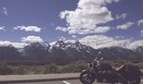 A scenic landscape features a mountain range in the background with snow-capped peaks under a partly cloudy sky. In the foreground, a motorcycle is parked on the side of the road, offering a sense of adventure and freedom.