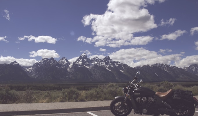 A scenic landscape features a mountain range in the background with snow-capped peaks under a partly cloudy sky. In the foreground, a motorcycle is parked on the side of the road, offering a sense of adventure and freedom.