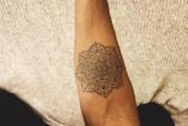 An arm with a detailed mandala tattoo is positioned against a textured fabric background. The tattoo features intricate geometric and floral patterns.
