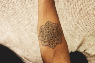 An arm with a detailed mandala tattoo is positioned against a textured fabric background. The tattoo features intricate geometric and floral patterns.