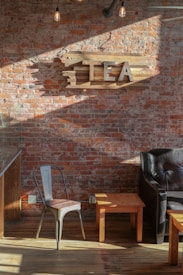 A rustic room with a brick wall featuring a wooden sign spelling 'TEA'. The space is furnished with a metal chair, wooden tables, and a dark leather chair. Exposed Edison bulbs hang from the ceiling, creating a cozy atmosphere with natural light casting shadows.