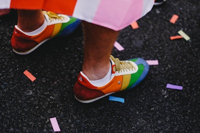 A pair of colorful sneakers featuring a rainbow pattern is worn by a person. The shoes are on a dark asphalt surface, adorned with scattered pieces of vibrant confetti in various colors, creating a festive and lively atmosphere.