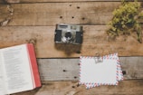 A vintage camera sits on a rustic wooden table next to an open book with a red cover. A bundle of dried yellow flowers is placed to the side, adding to the vintage aesthetic. Two envelopes with airmail design are clipped together next to the camera.